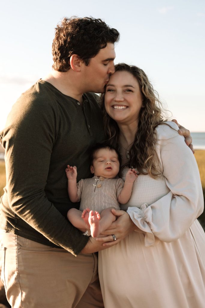 Brewster MA Newborn Beach Session | Brianna, Tyler & Baby Graham 4 Brewster MA Newborn Beach Session photo of a family with the dad kissing the mom's head on a Cape Cod beach